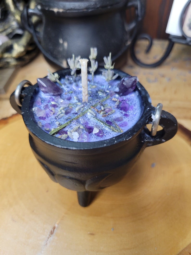 May include: A black cauldron-shaped candle with a purple wax interior. The candle is decorated with amethyst crystals, dried lavender, and a central wick. The cauldron has three small legs and two metal handles.