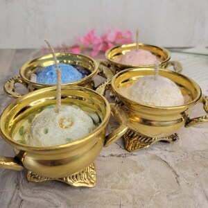 May include: Four gold-colored candle holders, each with a different colored candle inside: blue, pink, white, and green. The candles have wicks and are in ornate holders with handles and decorative bases. The background is a light gray and white.
