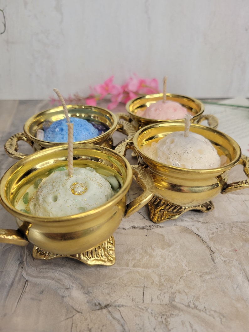 May include: Four gold-colored candle holders, each with a different colored candle inside: blue, pink, white, and green. The candles have wicks and are in ornate holders with handles and decorative bases. The background is a light gray and white.
