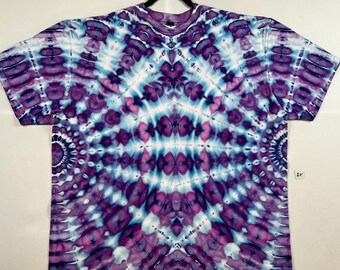 Puede incluir: Una camiseta teñida anudada en tonos morados, azules y blancos. La camiseta presenta un patrón simétrico con un diseño central que irradia hacia afuera. La tela es suave y la camiseta está colgada en una percha negra.