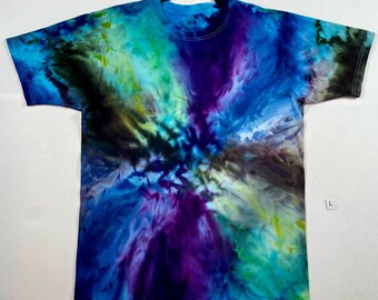 Puede incluir: Camiseta teñida con tinte anudado en tonos azules, morados, verdes y amarillos. La camiseta de cuello redondo presenta un patrón en espiral que irradia desde el centro. La camiseta de manga corta está colgada en una percha negra.