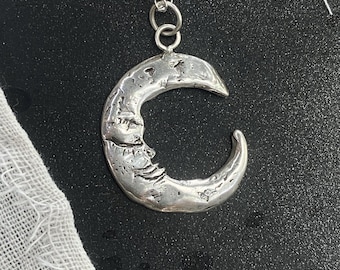 Puede incluir: Un collar con un colgante de media luna de plata con una superficie texturizada. La cadena es de plata y tiene un diseño de eslabones delicados.