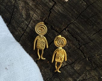 May include: A pair of gold-colored earrings. Each earring features a stylized human figure with a spiral head and outstretched arms and legs. The earrings are displayed on a dark, textured wooden surface.