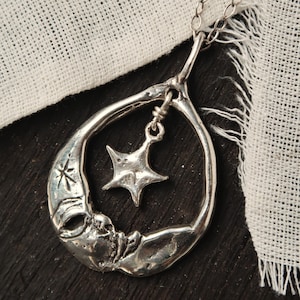 May include: A silver necklace with a crescent moon pendant and a dangling star charm. The pendant is textured and has a detailed design. The necklace is on a white fabric background.
