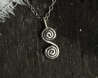 Può includere: Collana d'argento con ciondolo a doppia spirale su sfondo in legno scuro.