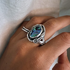Könnte beinhalten: Silberner Ring mit ovaler Abalone-Schale in schillernden Blau-, Grün- und Lilatönen. Der Ring hat eine detaillierte, verzierte Fassung mit Wirbeln und einem geformten Design am Ringband.