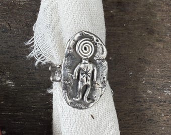 May include: A silver-toned ring featuring a stylized figure with a spiral head and star-like torso. The ring is set on a white fabric, showcasing its intricate design and unique aesthetic. The ring's design is reminiscent of ancient art.