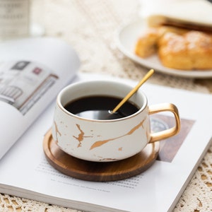 May include: A white ceramic coffee mug with gold marble pattern and a gold handle. The mug is filled with coffee and has a gold spoon inside. The mug is sitting on a wooden coaster on top of a magazine.