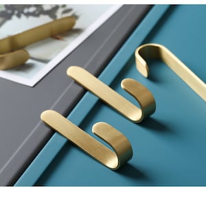 May include: Three gold-colored metal hooks with a simple, curved design. The hooks are on a blue surface.