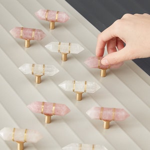 May include: Assortment of pink and white crystal cabinet knobs with gold accents. The knobs are shaped like a pointed crystal and have a gold metal base.