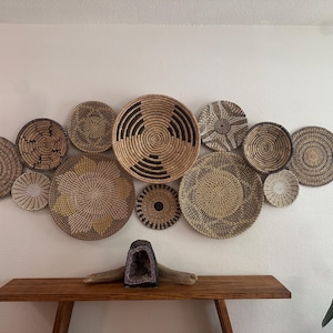 May include: A collection of twelve round, woven wall baskets in various sizes and patterns, featuring natural tan, brown, and black fibres. Below the baskets sits a dark purple amethyst geode resting on a piece of driftwood.