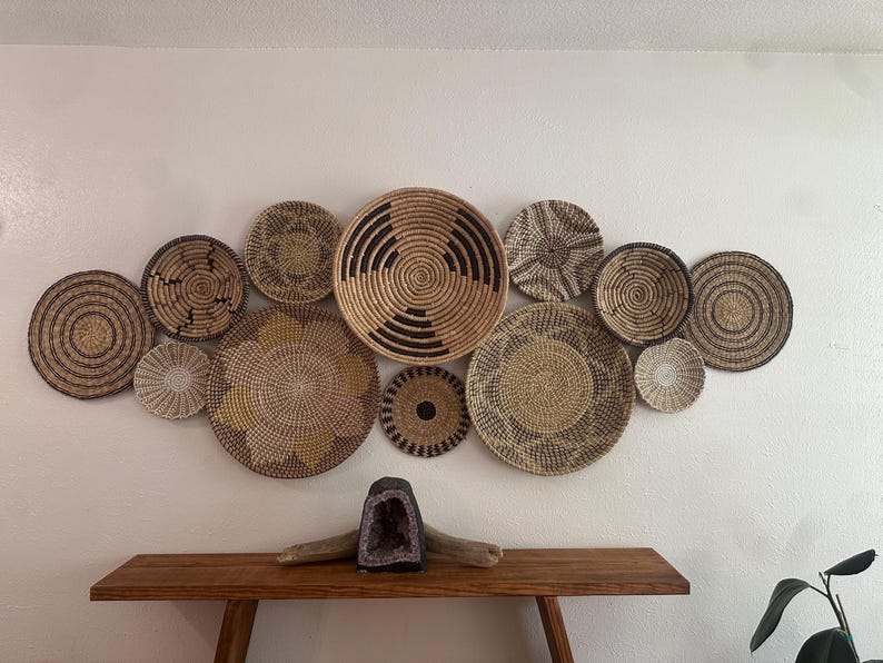 May include: A collection of twelve round, woven wall baskets in various sizes and patterns, featuring natural tan, brown, and black fibres. Below the baskets sits a dark purple amethyst geode resting on a piece of driftwood.