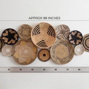 May include: A collection of twelve round woven wall baskets in natural tan, brown, and black tones with various geometric patterns. A metal ruler at the bottom indicates a total width of approximately 249 cm for the entire arrangement.