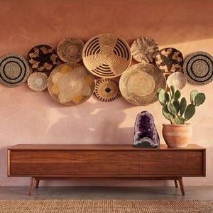 May include: A collection of round woven wall baskets in natural tan, brown, and black tones arranged in a cluster. A large purple amethyst geode and a green prickly pear cactus in a terracotta pot sit on a long, low wooden media console.