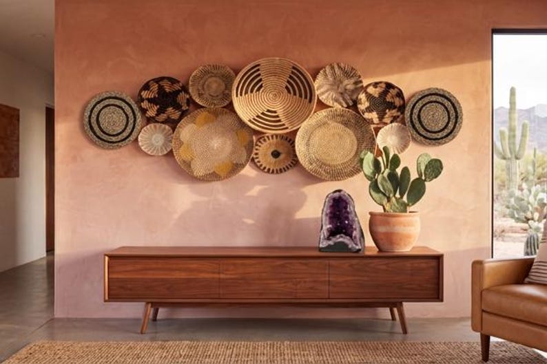May include: A collection of round woven wall baskets in natural tan, brown, and black tones arranged in a cluster. A large purple amethyst geode and a green prickly pear cactus in a terracotta pot sit on a long, low wooden media console.