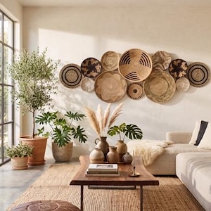 May include: A collection of twelve round woven wall baskets in various shades of tan, brown, and black arranged in a cluster on a light-coloured wall above a beige sofa, a wooden coffee table, and several potted green plants in a room.