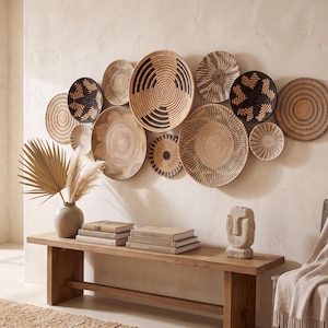 May include: A collection of twelve round woven wall baskets in varying sizes, featuring natural tan and black geometric patterns. The baskets are arranged in a cluster on a wall above a wooden bench, showcasing intricate coiled textures and designs.