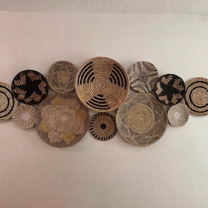 May include: A collection of twelve round woven wall baskets arranged in a cluster. The baskets feature various natural fibre textures and patterns in shades of tan, beige, brown, and black, including concentric circles, star motifs, and solid weaves.