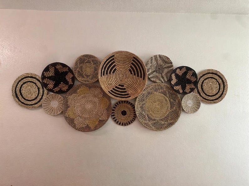 May include: A collection of twelve round woven wall baskets arranged in a cluster. The baskets feature various natural fibre textures and patterns in shades of tan, beige, brown, and black, including concentric circles, star motifs, and solid weaves.
