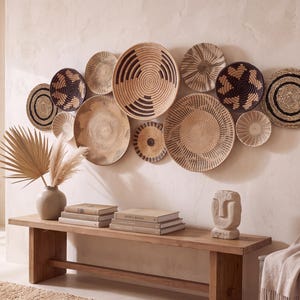 May include: A collection of twelve round woven wall baskets in varying sizes, featuring natural tan, beige, and dark brown geometric patterns. The baskets are arranged in a cluster on a wall above a wooden bench, displaying intricate coil textures.