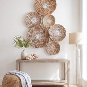 May include: A collection of six round woven wall baskets in natural tan and white, arranged in a cluster. The baskets feature intricate coiled patterns and varying sizes, mounted together to create a decorative display on a plain wall surface.