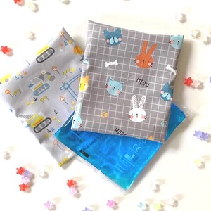 Könnte beinhalten: Zwei Stoff-Kältepackungen mit unterschiedlichen Designs. Eine Kältepackung hat einen grauen Hintergrund mit einem Muster aus Baufahrzeugen und dem Text "WOOF". Die andere Kältepackung hat einen grauen Hintergrund mit einem Gittermuster und Bildern von Katzen, Hasen und dem Text "MEOW".