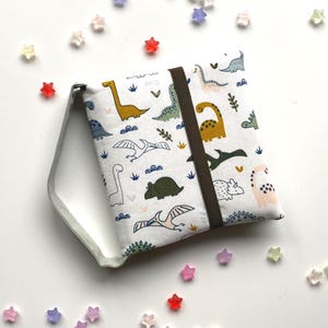 Könnte beinhalten: Eine weiße Stofftasche mit einem farbenfrohen Dinosaurier-Muster, das verschiedene Dinosaurier-Illustrationen in Grün, Gelb und Blau zeigt. Die Tasche hat ein braunes Gummiband und einen silbernen Metallgriff. Sterne-förmige Perlen sind verstreut.