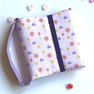 Könnte beinhalten: Ein lilafarbener Stoffbuchumschlag mit einem Blumenmuster mit rosa Blüten, gelben Stielen und lila Pilzen. Der Umschlag hat einen lilafarbenen Riemen mit einem schwarzen Gummiband.