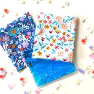 Könnte beinhalten: Zwei Stoffquadrate mit Blumenmustern, eines blau und eines hellblau, liegen übereinander auf einem blauen Gel-Kältepack. Das Kältepack hat ein weißes Etikett mit Text und Symbolen.