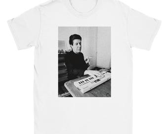 May include: White t-shirt featuring a black and white image of a person playing a keyboard. The person is wearing a dark-colored shirt and holding a cigarette. The keyboard is white with black keys.