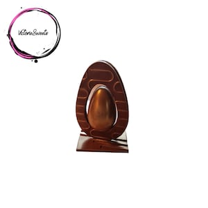 May include: A chocolate Easter egg with a decorative design, standing on a base. The egg is a dark brown colour and has a gold-coloured egg inside.