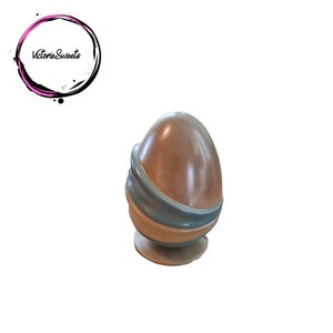May include: A light brown chocolate egg with a blue and white ribbon design. The egg is sitting on a small, round base.