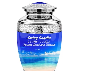 May include: A silver cremation urn with a blue band featuring a beach scene. The urn has decorative floral patterns and the text "Loving Angeila 2-3-1950 - 2-2-2022 Forever Loved and Missed". The lid is also blue.