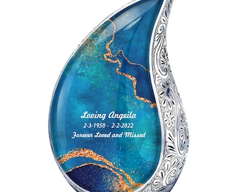 May include: A silver teardrop-shaped cremation urn with a blue and gold design. The urn has an inscription that reads "Loving Angeila 2-3-1950 - 2-2-2022 Forever Loved and Missed".
