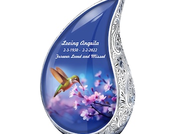 May include: A silver teardrop-shaped urn with a blue glass insert featuring a hummingbird and pink flowers. The urn is engraved with the text "Loving Angeila 2-3-1950 - 2-2-2022 Forever Loved and Missed".