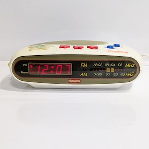 May include: A vintage Playskool alarm clock with a cream-colored body and red digital display showing 12:07. It has red and blue buttons, and an AM/FM radio dial. The clock is on a white surface.