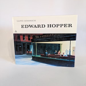 Puede incluir: Una portada de libro con una pintura de Edward Hopper titulada "Nighthawks". La pintura representa un restaurante de noche con tres clientes y un trabajador de mostrador. El restaurante está iluminado por letreros de neón y farolas. La portada del libro es blanca con texto negro que dice "LLOYD GOODRICH EDWARD HOPPER".