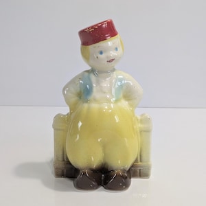 May include: A vintage ceramic figurine of a person wearing a red fez, blue vest, and yellow pants. The figurine has brown shoes and is seated on a small, decorative bench. The figure's arms are at their sides.