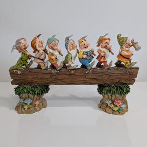 May include: A figurine of the Seven Dwarfs from Snow White, walking on a log. Each dwarf is dressed in a different outfit and carries a tool. The base is decorated with mushrooms and foliage, creating a whimsical scene.