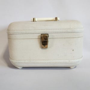 Puede incluir: Un neceser vintage de color crema con forma rectangular, bordes redondeados y un asa. El estuche presenta un cierre y adornos dorados, y pequeños pies. El asa es blanca. Es probable que el estuche esté hecho de un material texturizado.