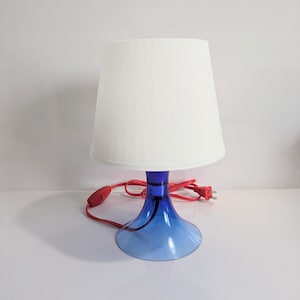 May include: A modern table lamp with a white lampshade and a blue base. The base has a flared design, and the lamp has a red power cord. The lamp is designed for indoor use and provides ambient lighting.