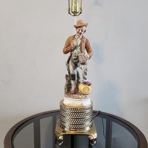 May include: A ceramic lamp featuring a figurine of a man holding a mug. The lamp base is gold with a black and white geometric pattern. The lamp is displayed on a round glass table with a wooden frame.