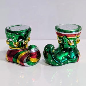 May include: A pair of Christmas-themed salt and pepper shakers shaped like elf boots. The boots are green, red, and gold with silver tops. The green sections have white polka dots and gold accents.