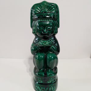 May include: A dark green ceramic figurine of a seated figure with a headdress. The figurine has a glossy finish and detailed features, including facial features and patterns. The base of the figurine has text. The design suggests a cultural or historical theme.