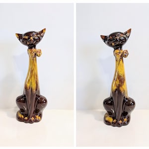 Op de afbeelding: Een keramieken kattenbeeldje met een lange nek en een strikje. De kat is voornamelijk bruin met gele en bruine accenten. Het beeldje heeft een glanzende afwerking en zit rechtop.