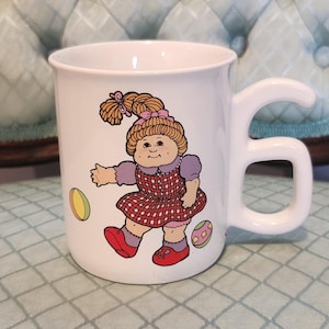 Könnte beinhalten: Weiße Keramiktasse mit einem Cartoon-Bild einer Cabbage Patch Kid-Puppe, die ein rot-weiß kariertes Kleid trägt und einen Ball hält. Die Tasse hat einen einzigartigen Henkel in Form eines Buchstabens 'C'.