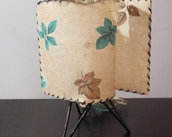 May include: A vintage table lamp with a white and brown floral patterned shade. The shade is made of a textured fabric and has a black trim. The lamp has a black metal base with three legs.