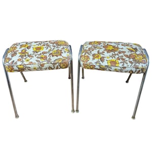 May include: Two vintage footstools with floral patterned cushions. The cushions feature a cream background with yellow and brown floral designs. The stools have chrome-colored metal frames and four legs each.