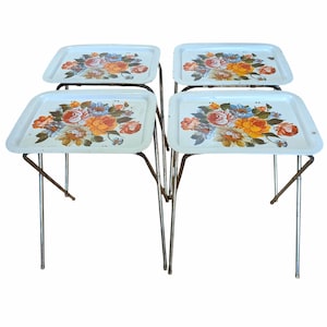 May include: A set of four light blue metal TV tray tables with folding legs. Each rectangular tray features a central floral pattern with orange, yellow, pink, and blue flowers and green leaves painted on the surface of the metal tops.