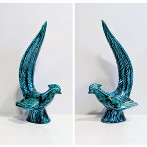 Op de afbeelding: Twee keramische vogelsculpturen in een turquoise glazuur. Elke vogel heeft een lange, omhoog gebogen staart en staat op een getextureerde basis. De sculpturen worden gepresenteerd tegen een witte achtergrond, die hun gedetailleerde ontwerp benadrukt.